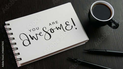 YOU ARE AWESOME! black lettering in notebook with cup of coffee and pens on black wooden desk