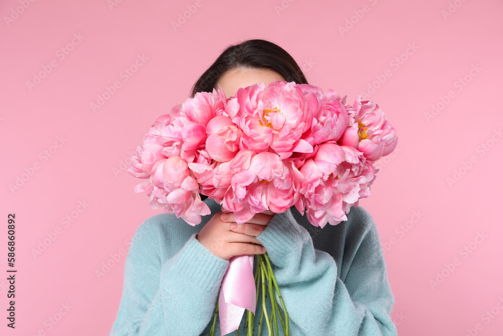 Young woman covering her face with bouquet of peonies on pink background