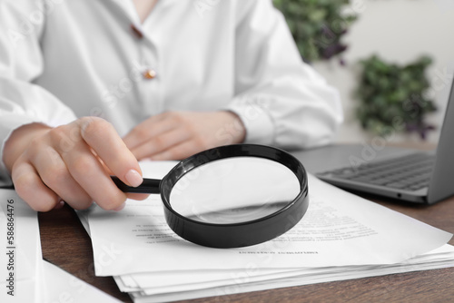 Woman looking at document through magnifier at wooden table indoors, closeup. Searching concept