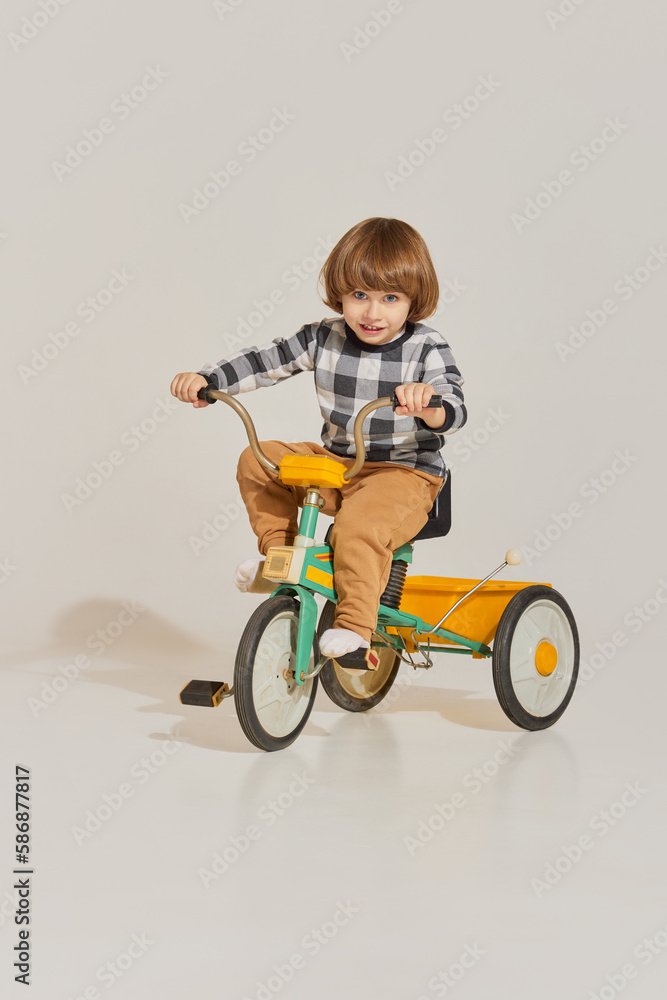 Charming cute boy with long hair riding retro bike and looking at camera over light background Charming cute boy with long hair riding retro bike and looking at camera over light background