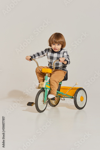 Charming cute boy with long hair riding retro bike and looking at camera over light background