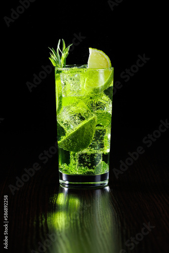Green tarragon lemonade with lime slices in a tall glass with ice on a black background.