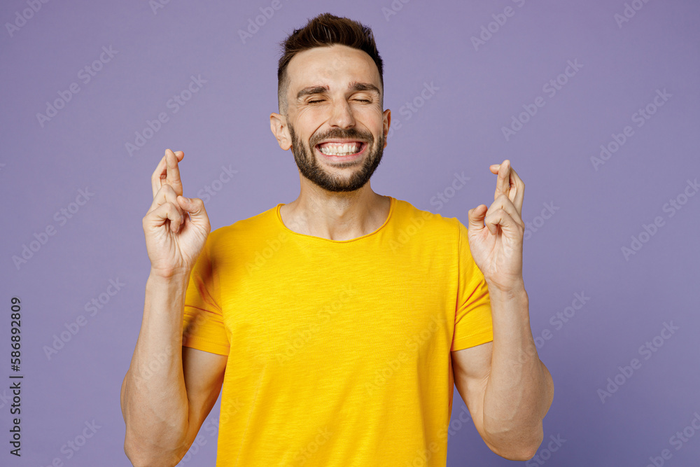 Young worried man wear yellow t-shirt waiting for special moment, keeping fingers crossed, making wish, eyes closed isolated on plain pastel light purple background studio portrait. Lifestyle concept. Young worried man wear yellow t-shirt waiting for special moment, keeping fingers crossed, making wish, eyes closed isolated on plain pastel light purple background studio portrait. Lifestyle concept.