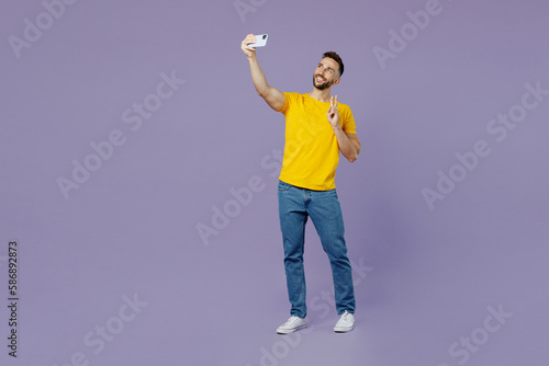 Full body young smiling happy caucasian man wear yellow t-shirt doing selfie shot on mobile cell phone post photo on social network show v-sign isolated on plain pastel light purple background studio. © ViDi Studio