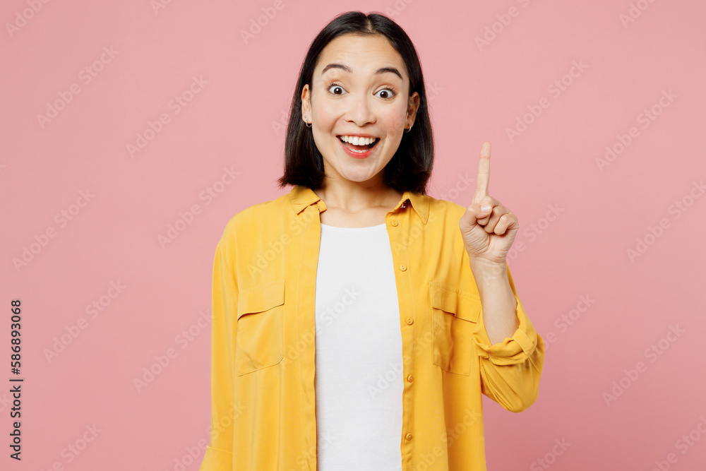Young fun insighted smart proactive woman of Asian ethnicity wear yellow shirt white t-shirt holding index finger up with great new idea isolated on plain pastel light pink background studio portrait. Young fun insighted smart proactive woman of Asian ethnicity wear yellow shirt white t-shirt holding index finger up with great new idea isolated on plain pastel light pink background studio portrait.