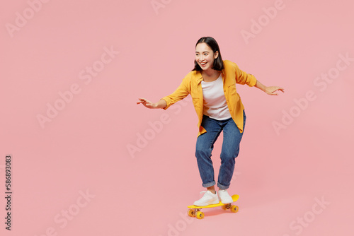 Full body young happy fun cool woman of Asian ethnicity wear yellow shirt white t-shirt riding skateboard pennyboard isolated on plain pastel light pink background studio portrait. Lifestyle concept.