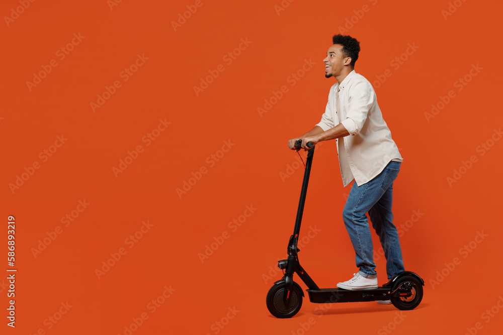Full body side view confident cool student young man of African American ethnicity wear light shirt casual clothes riding e-scooter isolated on orange red background studio portrait Lifestyle concept Full body side view confident cool student young man of African American ethnicity wear light shirt casual clothes riding e-scooter isolated on orange red background studio portrait Lifestyle concept