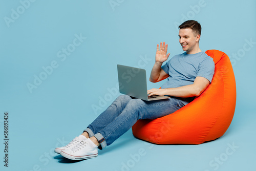 Full body young man wear casual t-shirt sit in bag chair hold use work on laptop pc computer waving hand get video call isolated on plain pastel light blue cyan background studio. Lifestyle concept.
