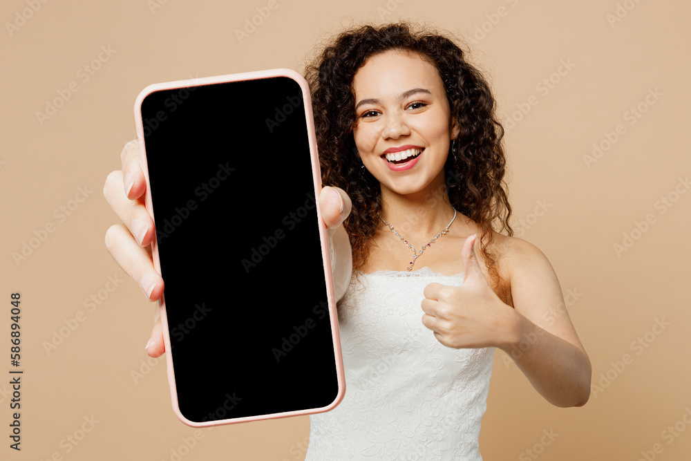 Obraz premium Happy young woman bride wear white wedding dress posing hold use mobile cell phone with blank screen workspace area show thumb up isolated on plain beige background Ceremony celebration party concept.