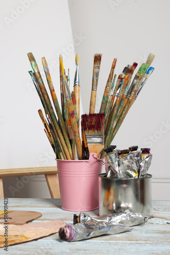 Easel with blank canvas and different art supplies on wooden table near white wall
