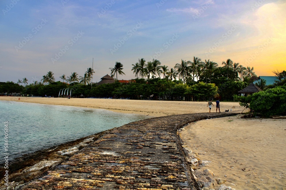 A couple walking on a beautiful landscape walkway between the ocean on the left and sandy beach on the right, overlooking a fiery sunset and palm trees. Shot in Bali, Indonesia.