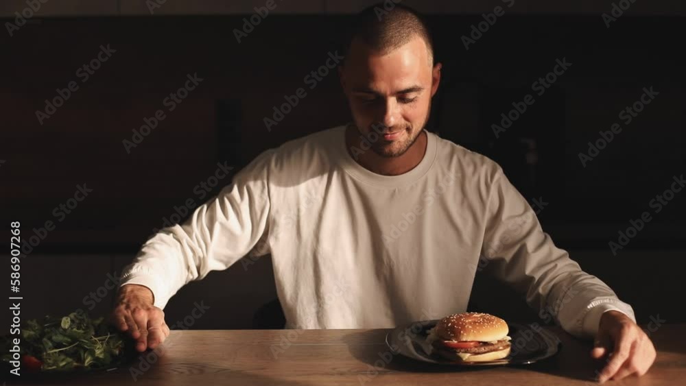 Man make choice salad or burger, unhealthy or healthy food, diet. Male look with aversion on salad and choose burger pushed the plate away. Male make decision eat unhealthy food, show thumb up. Man make choice salad or burger, unhealthy or healthy food, diet. Male look with aversion on salad and choose burger pushed the plate away. Male make decision eat unhealthy food, show thumb up.