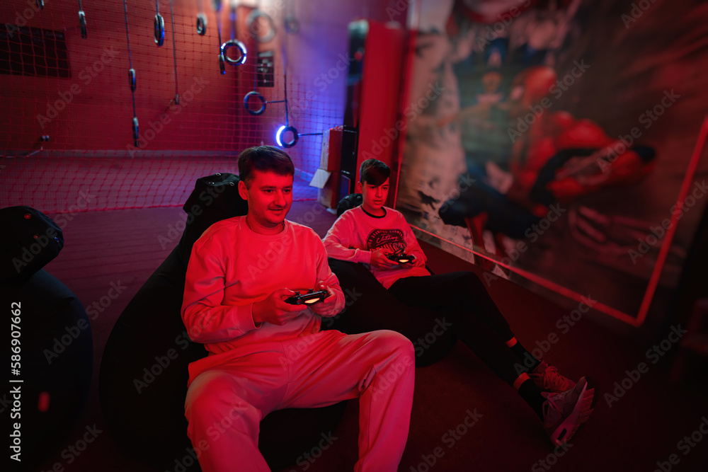 Father and son play gamepad video game console in red gaming room. Dad and kid gamers.