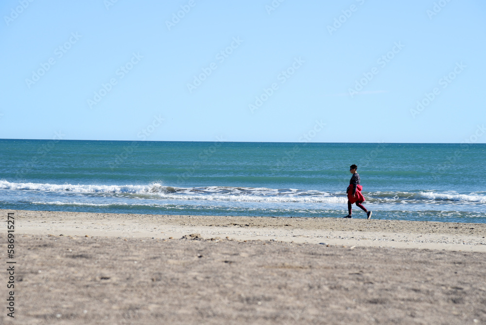 Girl walk along the coastline of the sea in autumn. Woman walk barefoot on beach near sea. Persons walking on empty beach in winter season. Sea beach. Tourist travels in the mediterranean.