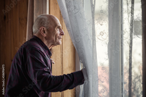 Single elderly man looking through window glass standing at home. Grandpa holding white curtain with hand and look out view from window.