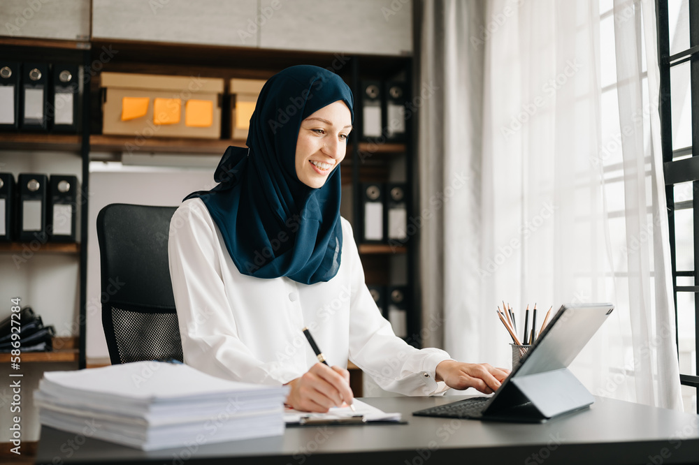 Young Muslim woman working with laptop and tablet in her modern business home office.. Young Muslim woman working with laptop and tablet in her modern business home office..