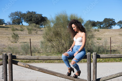 curly-haired, brunette, Spanish woman resting on the fence of the path leading to the meadow. She is dressed in jeans and a white top and enjoys the sunny day. The woman is sad and depressed.