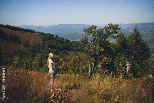 A slender young girl with long blond hair, in summer clothes, stands in the mountains, on a hot sunny day, and looks into the distance.