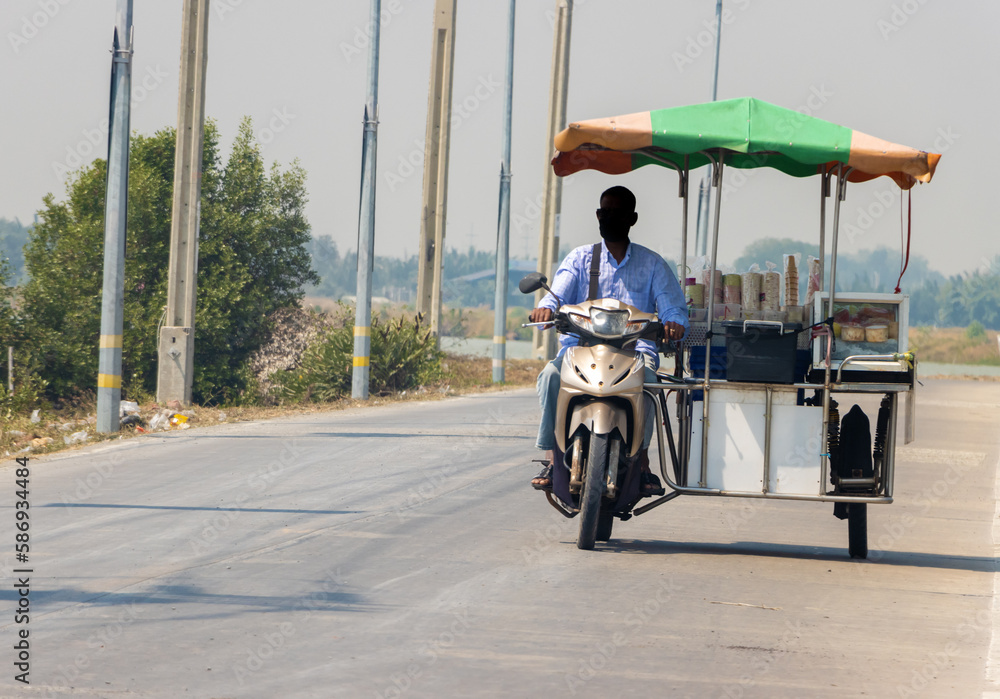 Obraz premium An ice cream vendor rides a three-wheeled motorcycle along a village road, Thailand
