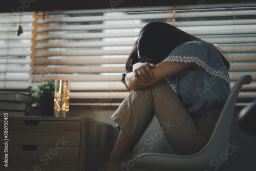 Sad and depressed young woman sitting nere window in the living room, looking outside with a sad expression, conveying feelings of exhaustion, loneliness, and unhappiness. Vintage style photo