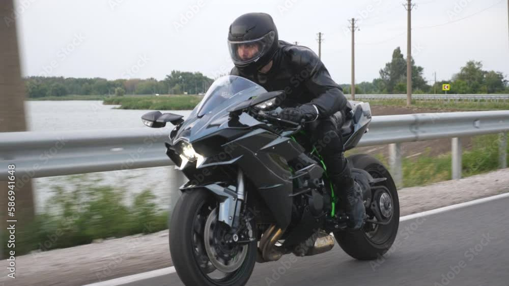 Motorcyclist races his motorcycle on country road near river or lake. Man in helmet rides fast on modern sport motorbike at rural route. Guy driving bike during trip. Concept of freedom and journey Motorcyclist races his motorcycle on country road near river or lake. Man in helmet rides fast on modern sport motorbike at rural route. Guy driving bike during trip. Concept of freedom and journey