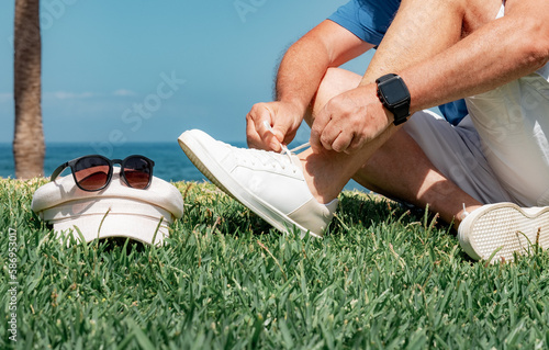 Close up Shot of Caucasian Elderly Man Tying his Shoe Laces. Mature Man in Outdoor at Sea in Sunny Day Sitting on Green Meadow