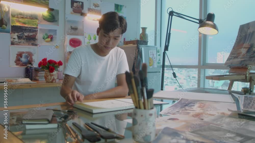 Young Asian man walking in art studio, sitting at desk and using pencil while drawing sketch in album Young Asian man walking in art studio, sitting at desk and using pencil while drawing sketch in album