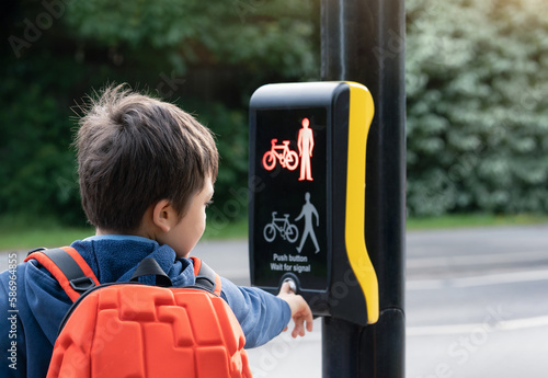 Rear view portrait School kid pressing  button at traffic lights on pedestrian crossing on way to school.Child boy with backpack using traffic signal controlled pedestrian facilities for crossing road photo