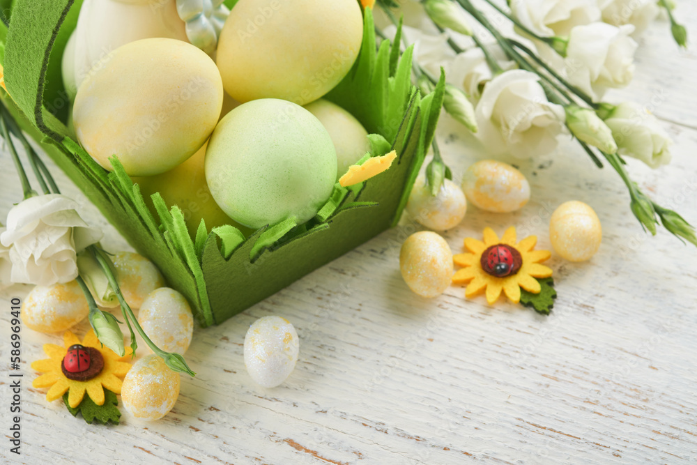 Happy Easter. Easter eggs and rabbit in green basket on white old wooden table with white and yellow roses. Spring Happy Easter holiday card.  Easter background with copy space. Top view.