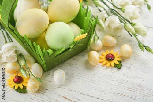 Happy Easter. Easter eggs and rabbit in green basket on white old wooden table with white and yellow roses. Spring Happy Easter holiday card. Easter background with copy space. Top view.