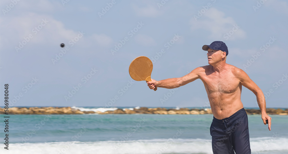 A handsome mature man hits a ball while playing matkot on the beach