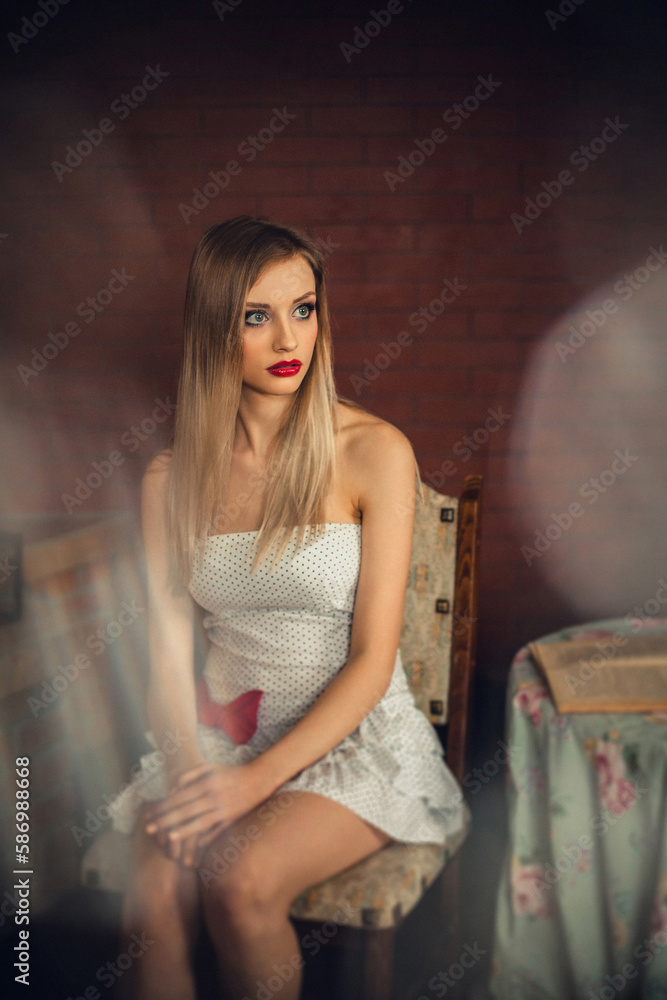 A gorgeous girl with bright make-up, with long blond hair, in a light dress, sits in a room at a table.