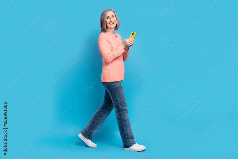 Full length cadre of excited old business lady walk steps hold smartphone browsing sliding shopping web site online isolated on blue color background Full length cadre of excited old business lady walk steps hold smartphone browsing sliding shopping web site online isolated on blue color background