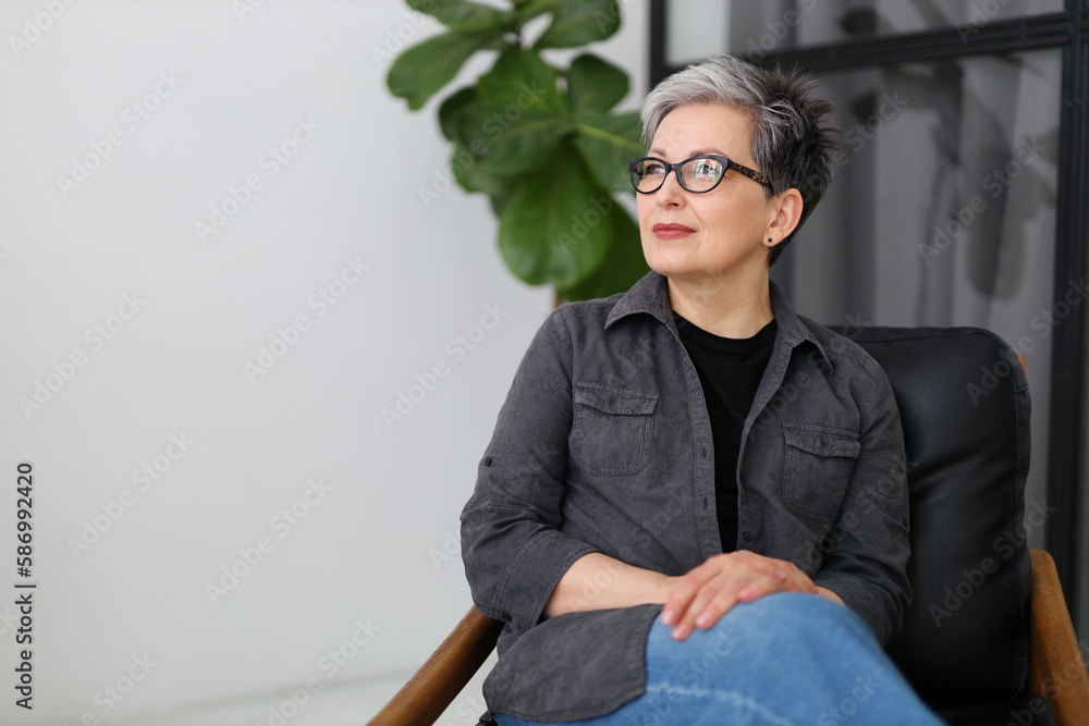 Mature pensive businesswoman in glasses and black clothes sits on an armchair in the office, empty space for text. Mature pensive businesswoman in glasses and black clothes sits on an armchair in the office, empty space for text.