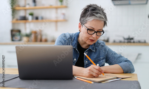 An adult woman writes in a notebook at a table with a laptop. Mature entrepreneur.
