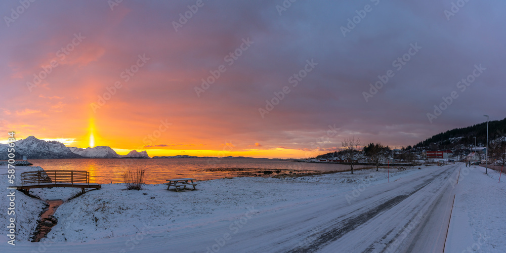 Abendrot mit einem hellen Sonnenstrahl über den verschneiten Bergen von Senja, Norwegen. die roten Wolken spiegeln sich in Wasser des Fjords bei Skaland. Sonnenuntergang am Bergsfjorden, Brücke