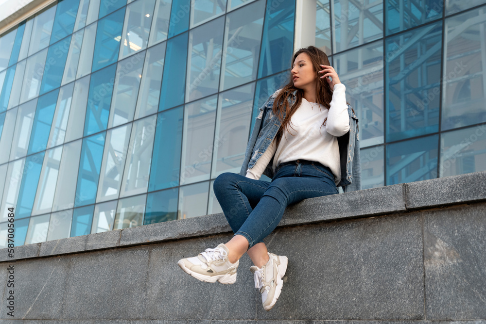 Stylish young woman resting on summer city street. Beautiful student girl in jeans sitting near modern building