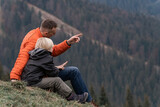 Father and son sit on hillside on the forest background. Trekking and hiking with children. Weekend hike in mountains.