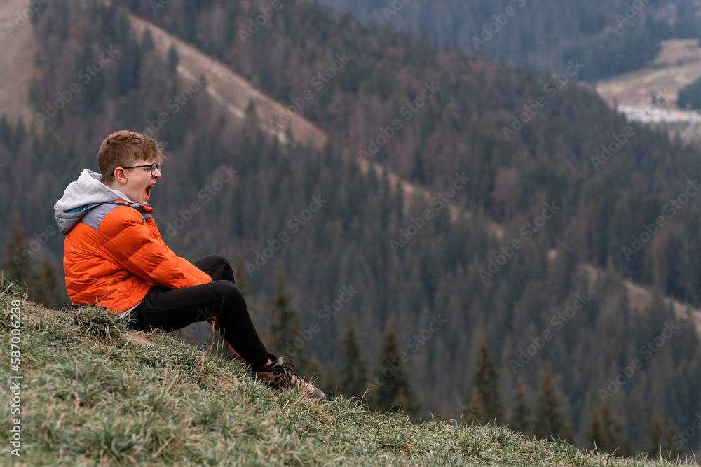 Bored guy sits on hillside and yawns. Boy screams in mountains. Portrait of child in the autumn on hike.