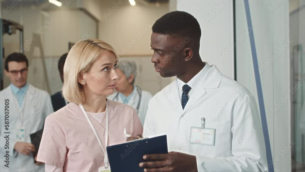 African American male doctor and Caucasian female nurse discussing patients test results standing at modern hospital corridor African American male doctor and Caucasian female nurse discussing patients test results standing at modern hospital corridor