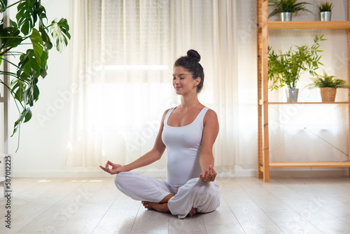 Pregnant american woman in leggings and crop top sitting on floor in living-room in lotus posture with closed eyes, doing mudra sign with fingers