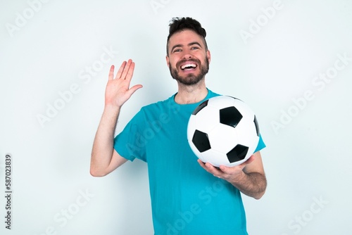 Young man holding a ball over white background Waiving saying hello happy and smiling, friendly welcome gesture. © Jihan
