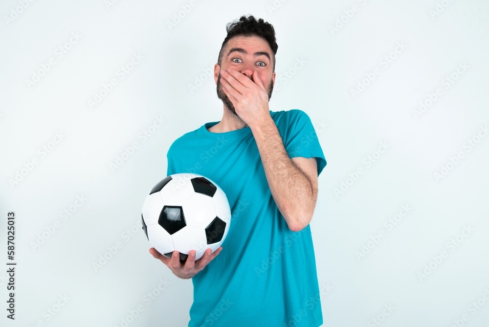 Vivacious Young man holding a ball over white background , giggles joyfully, covers mouth, has natural laughter, hears positive story or funny anecdote