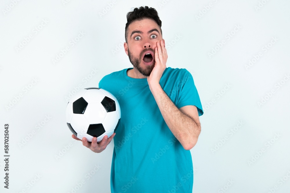 Stupefied Young man holding a ball over white background expresses excitement and thrill, keeps jaw dropped, hands on cheeks, has eyes popped out