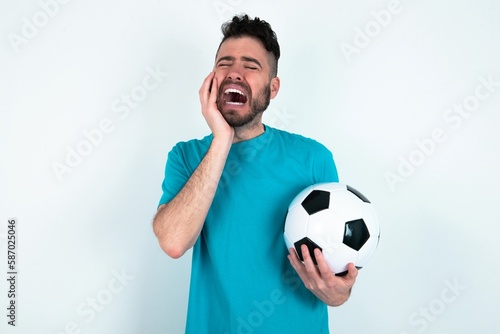 Doleful desperate crying Young man holding a ball over white background , looks stressfully, frowns face, feels lonely and anxious