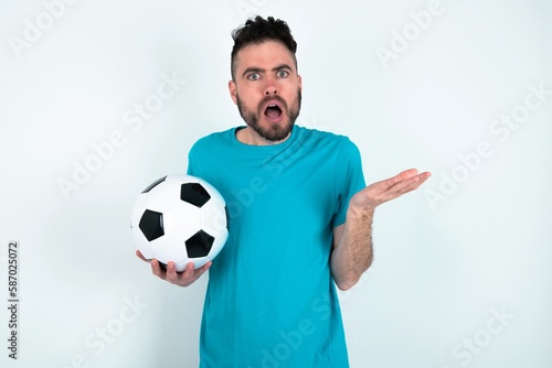 Frustrated Young man holding a ball over white background feels puzzled and hesitant, shrugs shoulders in bewilderment, keeps mouth widely opened, doesn't know what to do. © Jihan