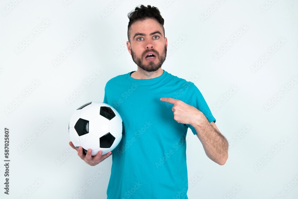 Embarrassed Young man holding a ball over white background indicates at herself with puzzled expression, being shocked to be chosen to participate in competition, hesitates about something