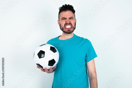 Young man holding a ball over white background keeps teeth clenched, frowns face in dissatisfaction, irritated because of much duties.