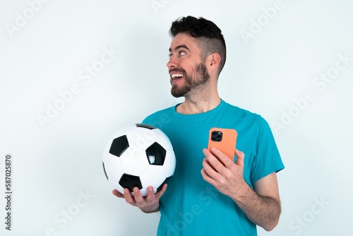 Young man holding a ball over white background holding a smartphone and looking sideways at blank copyspace.