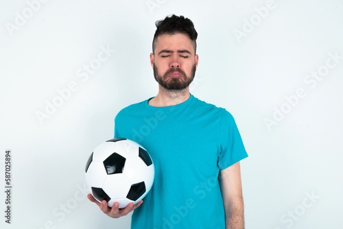 Dismal gloomy rejected Young man holding a ball over white background has problems and difficulties, curves lower lip and closes eyes in despair, being in depression